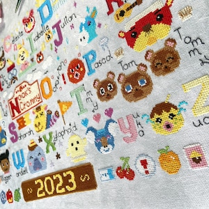 May include: Cross-stitch sampler featuring colorful cartoon characters and letters of the alphabet. The design includes the text "Nook's Cranny" and the year "2023". The sampler is on a light gray fabric.