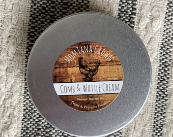 May include: A silver tin with a label that reads "Montana Grown Comb & Wattle Cream" with a chicken illustration. The label also says "Beeswax, essential oils Treats & Prevents Frostbite".