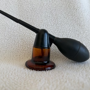 May include: A vintage amber glass perfume atomizer with a black bulb and nozzle. The atomizer has a rounded base and a black, cone-shaped top. The bulb is a dark, matte black. The nozzle extends at an angle from the top.