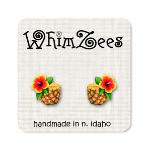 May include: A pair of pineapple earrings with a red and yellow hibiscus flower on top. The earrings are handmade in Idaho and feature the text "Whimzees".