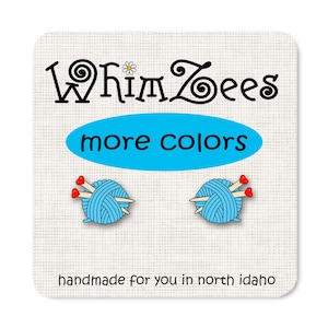 Op de afbeelding: Een paar blauwe garenbal-oorbellen met rode hartvormige breinaalden. De oorbellen zijn handgemaakt en hebben de tekst "Whimzees more colors" en "handmade for you in north idaho".