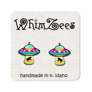 May include: A pair of whimsical earrings featuring a rainbow-colored UFO with a black cow inside. The earrings are handmade in Idaho and feature the text "WhimZeEs" on the card.