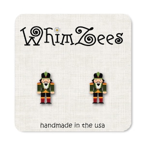 May include: A pair of WhimZee's stud earrings featuring a nutcracker soldier design. The earrings are on a white card with the brand name and the text "handmade in the usa". The nutcrackers are green, black, red, and gold.