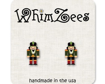 May include: A pair of WhimZee's stud earrings featuring a nutcracker soldier design. The earrings are on a white card with the brand name and the text "handmade in the usa". The nutcrackers are green, black, red, and gold.