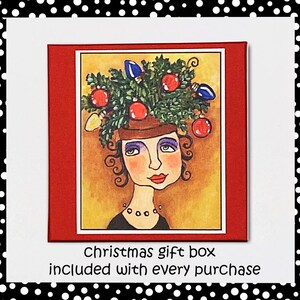 May include: A whimsical illustration of a woman wearing a Christmas tree on her head. The tree is decorated with red and blue ornaments and lights. The woman has brown hair, blue eyes, and a red mouth. The background is a yellow-gold color. The text "Christmas gift box included with every purchase" is printed below the illustration.