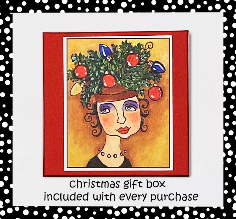 May include: A whimsical illustration of a woman wearing a Christmas tree on her head. The tree is decorated with red and blue ornaments and lights. The woman has brown hair, blue eyes, and a red mouth. The background is a yellow-gold color. The text "Christmas gift box included with every purchase" is printed below the illustration.
