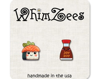 Könnte beinhalten: Ein Paar Emaille-Anstecknadeln auf einer weißen Karte mit der Aufschrift WhimZees und handmade in the usa. Eine Nadel zeigt ein Lachs-Sushi mit lächelndem Gesicht, die andere eine braune Sojasaucenflasche mit Gesicht und dem Aufdruck joy soy.