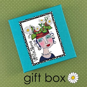 May include: A turquoise blue gift box with a white label featuring a whimsical illustration of a person with flowers growing out of their head. The label has a black border and the text "My Turn Toes" in a stylized font.