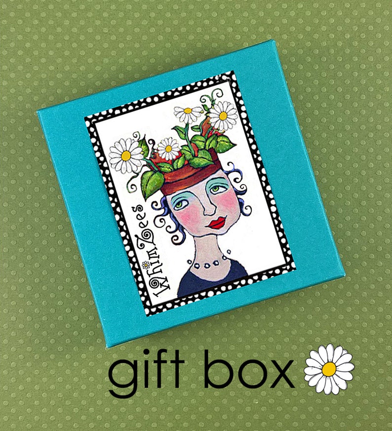 May include: A turquoise blue gift box with a white label featuring a whimsical illustration of a person with flowers growing out of their head. The label has a black border and the text "My Turn Toes" in a stylized font.