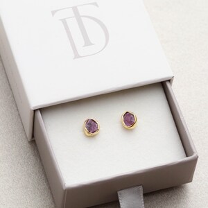 May include: A pair of gold stud earrings with small, irregular purple gemstones set in the center of each earring. The earrings are displayed in a small, white box with the letter "TD" embossed on the top.