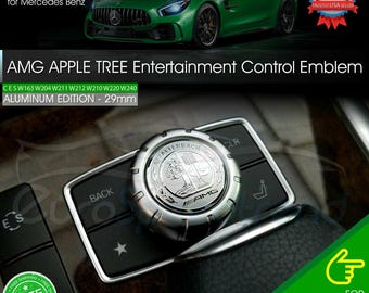 Puede incluir: Emblema de control de entretenimiento AMG Apple Tree plateado, 29 mm, para Mercedes Benz. El emblema es de aluminio y presenta el logotipo AMG y el texto "ASFALTEREACH". La imagen también muestra un coche Mercedes Benz verde.