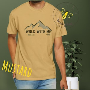 May include: Mustard-colored t-shirt with the text "WALK WITH ME" above a mountain graphic. The text "MOSES 6:34 -GOD" is below the mountain design. A "busy bee" logo is in the upper right corner.