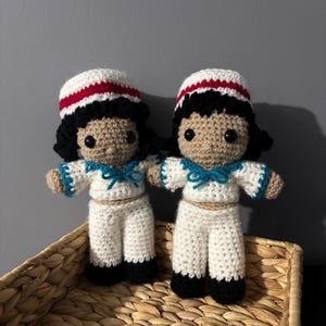 Peut inclure: Deux poupées en crochet faites à la main avec une peau bronzée, des cheveux noirs et des tenues de marin blanches avec des bordures bleues et des chapeaux à rayures rouges. Chaque poupée a des yeux en boutons noirs et des détails en laine.