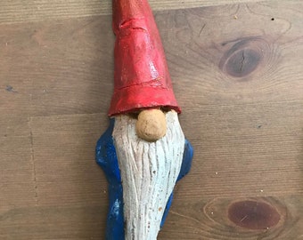 May include: A wooden gnome figurine with a red hat, white beard, and blue painted details. The gnome is carved from wood and painted in a rustic style.