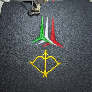 May include: A black fabric with a white embroidery hoop. The hoop contains an embroidered design of three arrows in the colours of the Italian flag, with a yellow bow and arrow underneath.