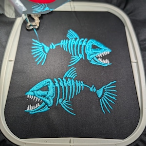 Könnte beinhalten: Zwei türkisfarbene Fischskelett-Stickereien auf schwarzem Stoff. Die Fischskelette haben detaillierte Rippen, Flossen und offene Münder mit Zähnen. Die Stickerei ist von einem weißen Stickrahmen umgeben.