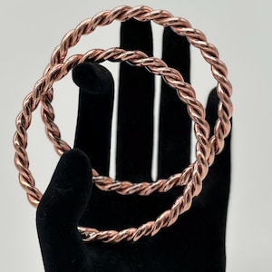 May include: Two copper-colored, twisted metal bangles. The bangles are circular and have a rope-like texture. The jewelry is displayed on a black velvet hand-shaped display stand against a white background.