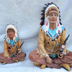 May include: Two Native American figurines, both wearing traditional headdresses and clothing, are seated with their legs crossed. They are holding pipes in their hands. The figurines are made of a light brown material and have painted details.