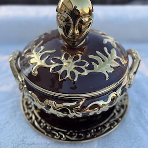 Peut inclure: Un bol de service en céramique brun avec un couvercle et une base dorés. Le couvercle présente un visage humain stylisé avec des accents dorés. Le bol et la base sont décorés de motifs floraux dorés.
