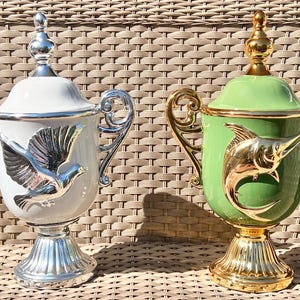 Pode incluir: Duas urnas decorativas com tampas, alças e bases de pedestal. Uma é branca com detalhes prateados e um desenho de pomba. A outra é verde com detalhes dourados e um desenho de peixe. Ambas são ornamentadas.