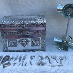 May include: A silver metal box with an ornate design featuring elephants and floral patterns. The box is closed and has a hinged lid. There are also several silver metal bells on a stand.