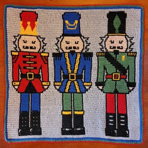 May include: Crocheted square featuring three colorful nutcracker figures. The figures are in red, blue, and green uniforms with black boots. The background is light gray, and the border is blue, red, and green.