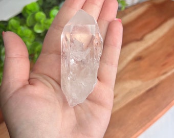 May include: A clear quartz crystal point with a natural, rough texture. The crystal is held in a hand, showcasing its size and shape.