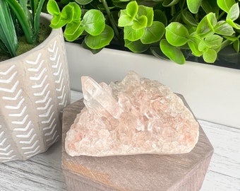 May include: A cluster of peach-colored quartz crystals sits atop a hexagonal wooden block. The crystals have a rough, textured surface and a pointed tip. The block is a light brown color with a wood grain pattern. Green plants are in the background.