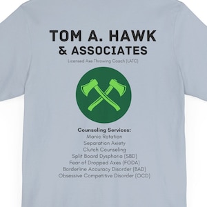 Puede incluir: Una camiseta azul claro con un logotipo circular verde que presenta dos hachas cruzadas. El texto "TOM A. HAWK & ASSOCIATES" está impreso sobre el logotipo. Debajo del logotipo, el texto "Licensed Axe Throwing Coach (LATC)" está impreso. Debajo del texto, se imprime una lista de servicios de asesoramiento: "Manic Rotation, Separation Anxiety, Clutch Counseling, Split Board Dysphoria (SBD), Fear of Dropped Axes (FODA), Borderline Accuracy Disorder (BAD), Obsessive Competitive Disorder (OCD)"