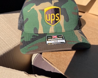 May include: A camouflage trucker hat with a black mesh back. The hat features a brown UPS logo with a yellow outline on the front. The hat is made by Richardson and has a size tag that says "Original 112 Trucker Adjustable".