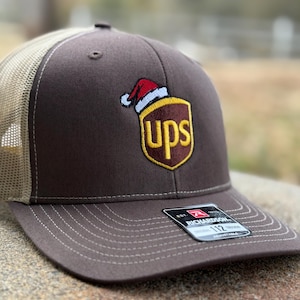 May include: Brown and tan trucker hat with a brown UPS logo with a Santa hat on top. The hat has a Richardson 112 tag on the side.
