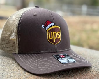 Op de afbeelding: Bruine en beige trucker cap met een bruin UPS-logo met een kerstmanmuts erop. De pet heeft een Richardson 112-label aan de zijkant.