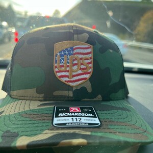 以下が含まれることがあります： 赤、白、青のアメリカ国旗をあしらった、茶色と金のUPSロゴが付いた迷彩柄のトラッカーハット。帽子は調節可能で、メッシュの黒い裏地が付いています。帽子には、「EST. 1970 RICHARDSON ORIGINAL 112 TRUCKER ADJUSTABLE」という文字が書かれた黒いラベルが付いています。