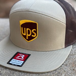 Puede incluir: Una gorra beige y marrón de visera plana y cierre a presión con una parte trasera de malla marrón. La gorra tiene un logotipo UPS bordado en marrón y amarillo en la parte delantera.