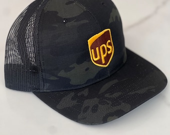 Op de afbeelding: Een zwarte en groene camouflage trucker cap met een bruin en geel UPS-logo geborduurd op de voorkant.
