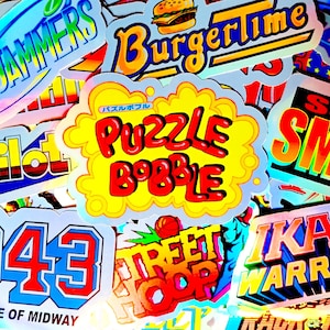 Könnte beinhalten: Eine Sammlung bunter, holografischer Vinyl-Aufkleber mit verschiedenen Logos und Titeln von Retro-Arcade-Spielen, darunter Puzzle Bobble, BurgerTime, 1943 The Battle of Midway, Street Hoop, Super Smash und Ikari Warriors, die dicht übereinander liegen.