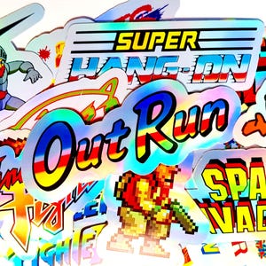 Op de afbeelding: Een verzameling holografische stickers met retro videogame-afbeeldingen. Ontwerpen zijn onder meer "Out Run" en "Space Invaders", evenals pixelpersonages. De stickers hebben een glanzende, iriserende afwerking en levendige kleuren.