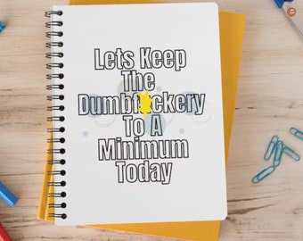 May include: A white spiral-bound notebook with the text "Lets Keep The Dumbfuckery To A Minimum Today" in black. The notebook is on top of a yellow notebook. Blue paper clips and scissors are visible.
