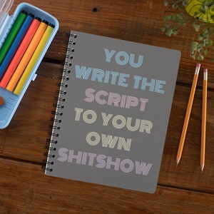 May include: A gray spiral bound notebook with the text "YOU WRITE THE SCRIPT TO YOUR OWN SHITSHOW" in colorful, striped lettering. The notebook is on a wooden surface with a blue pencil case and two yellow pencils.