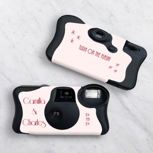 May include: A white and black disposable camera with the text "Turn on the flash!" and stars printed on the front. The camera also has the text "Camilla & Charles" printed on the front.