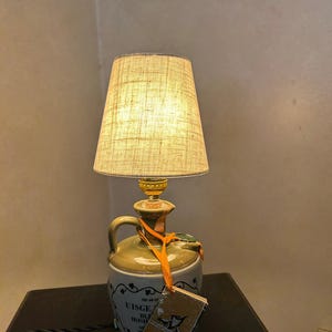 May include: A distinctive lamp featuring a beige fabric shade. The base is a ceramic jug with the words "Uisge Beatha Irish Whiskey" and a tag. The lamp has a handle and a decorative top.