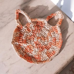 May include: A handmade, circular crochet coaster in the shape of a cat's face. The coaster features a spiral design with alternating orange and beige yarn, and two small handles. The coaster is approximately 6 inches in diameter.