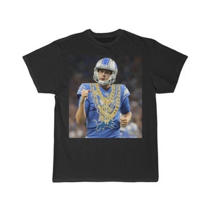 May include: A black t-shirt featuring a photograph of a football player wearing a blue jersey and a gold chain with dollar signs. The player is wearing a helmet and celebrating a touchdown.