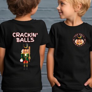 Könnte beinhalten: Zwei Kinder tragen schwarze T-Shirts mit einem Nussknacker-Motiv. Ein Shirt sagt "Crackin' Balls" und das andere sagt "The Ball Cracker".
