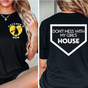 Könnte beinhalten: Schwarzes T-Shirt mit einem weißen Baseball-Diamanten-Motiv auf der Rückseite, auf dem "Don't mess with my girl's house" steht. Die Vorderseite des Shirts hat einen gelben Softball mit einer Catcher-Silhouette darin und den Text "Catcher's Mom".