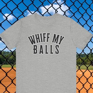 Puede incluir: Una camiseta gris jaspeado con el texto "WHIFF MY BALLS" en blanco y negro. La camiseta se muestra frente a un campo de béisbol, con una valla de alambre en primer plano.