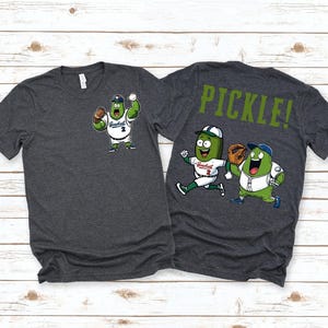 May include: Two dark gray t-shirts with a pickle baseball player graphic. The front shirt has a single pickle player with a baseball glove. The back shirt has two pickle players running with the text "PICKLE!" on the back.