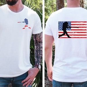 May include: White t-shirt with a baseball player silhouette holding a bat in front of an American flag on the front. The back of the shirt features a distressed American flag with a baseball player silhouette holding a bat.