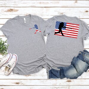 May include: Two grey t-shirts with a baseball player silhouette design. The silhouette is holding a baseball bat and is superimposed on an American flag. One shirt has the design on the front and the other on the back.