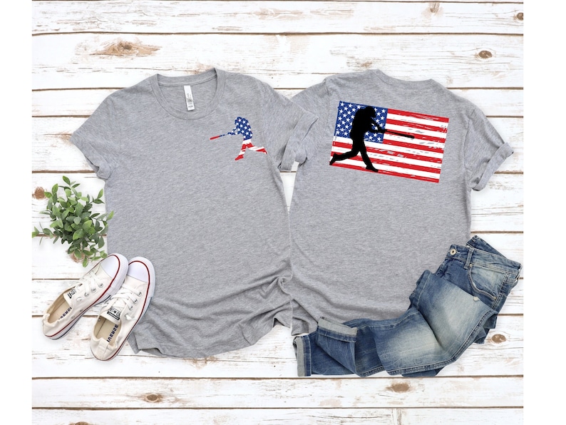 May include: Two grey t-shirts with a baseball player silhouette design. The silhouette is holding a baseball bat and is superimposed on an American flag. One shirt has the design on the front and the other on the back.
